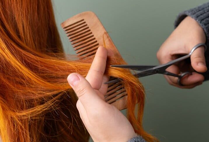Close-up of red hair being trimmed with scissors and comb on green background.